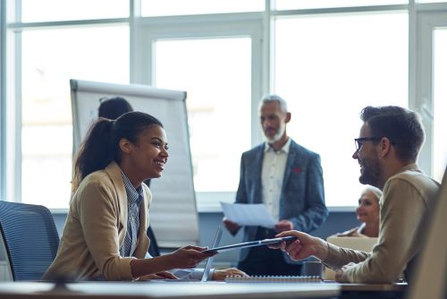 Two cheerful diverse office workers discussing something and smiling while having a meeting with colleagues in the modern office. People communicating at work